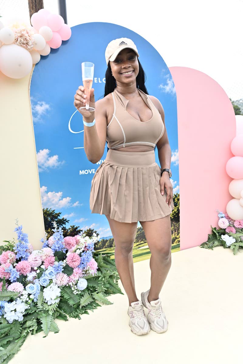 Content creator Sue-Tanya McHorgh raises a glass in a chic taupe activewear set featuring a fitted cropped top and pleated skirt, paired with a coordinating cap and sneakers.